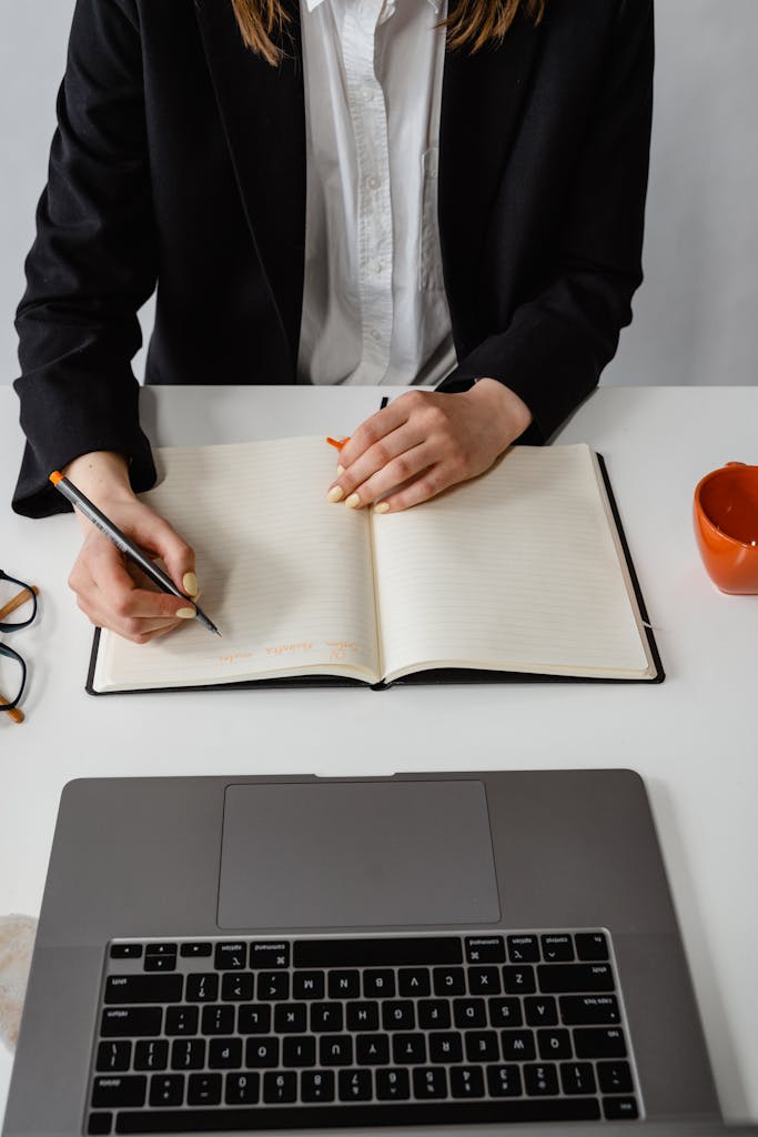 Professional woman writing in a notebook at her office desk, with a laptop in front.